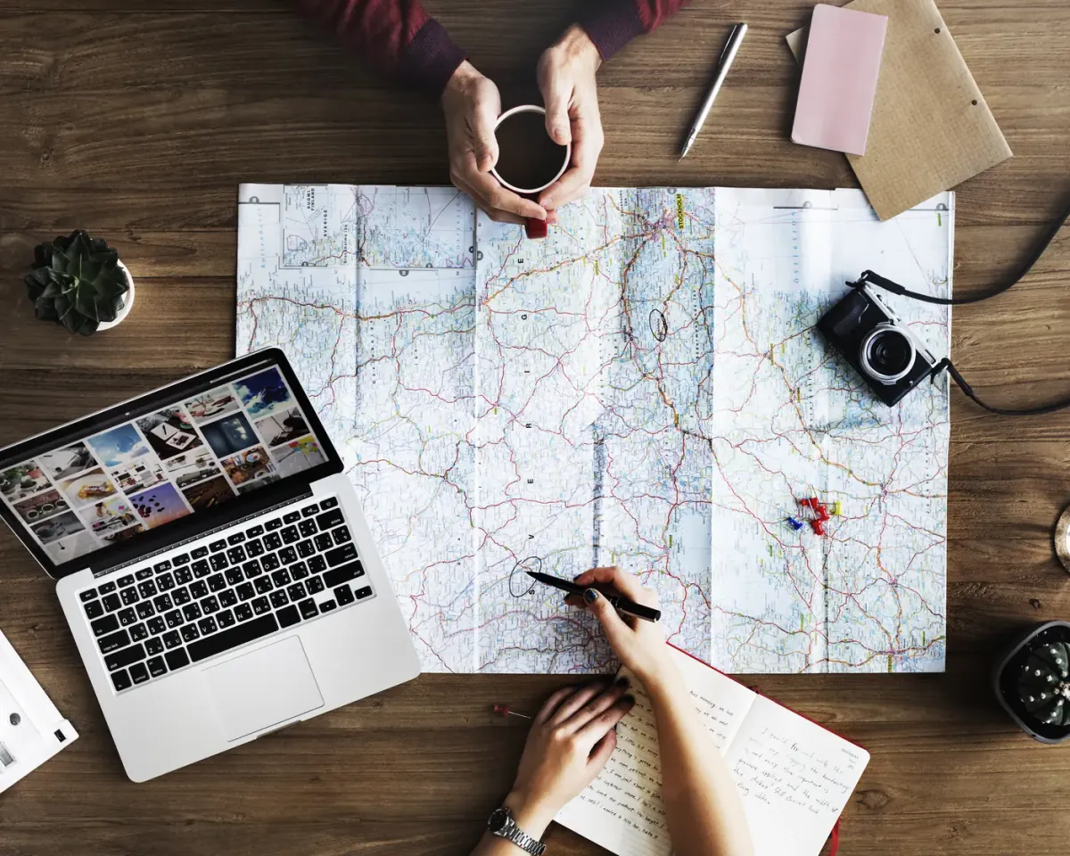 Dos personas planifican una ruta sobre un mapa extendido en una mesa de madera, con un café y un portátil al lado.